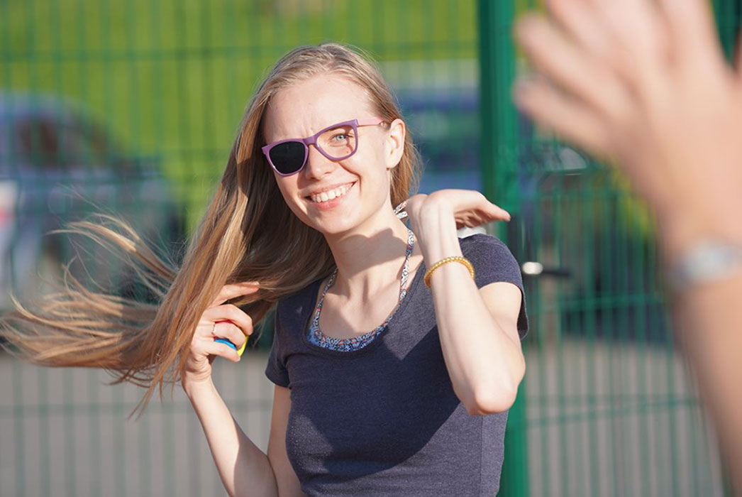 Girl in the sun wearing patch glasses so her left eye can see. Long hair blowing in the wind, and big smile